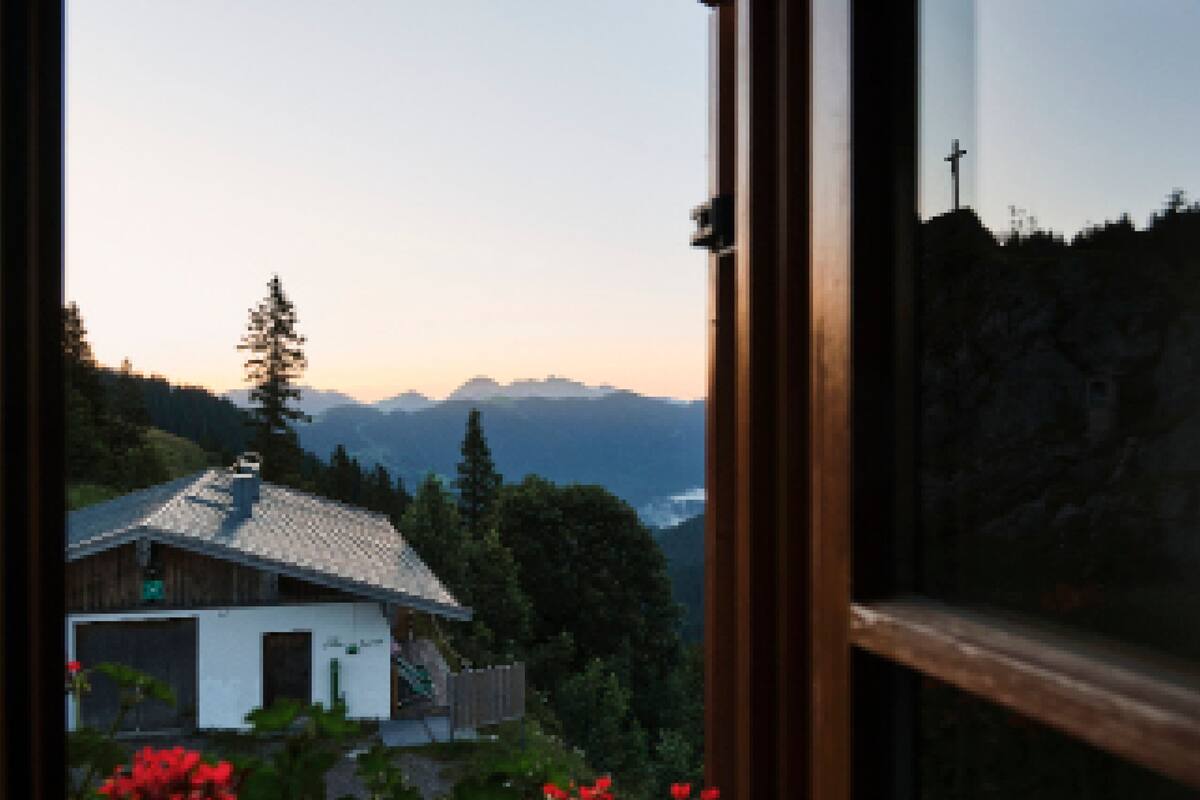 A view through a window of a house with a sloped roof, surrounded by greenery and mountains in the background.