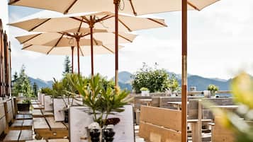 Outdoor seating with wooden tables and beige umbrellas, overlooking a mountainous landscape.