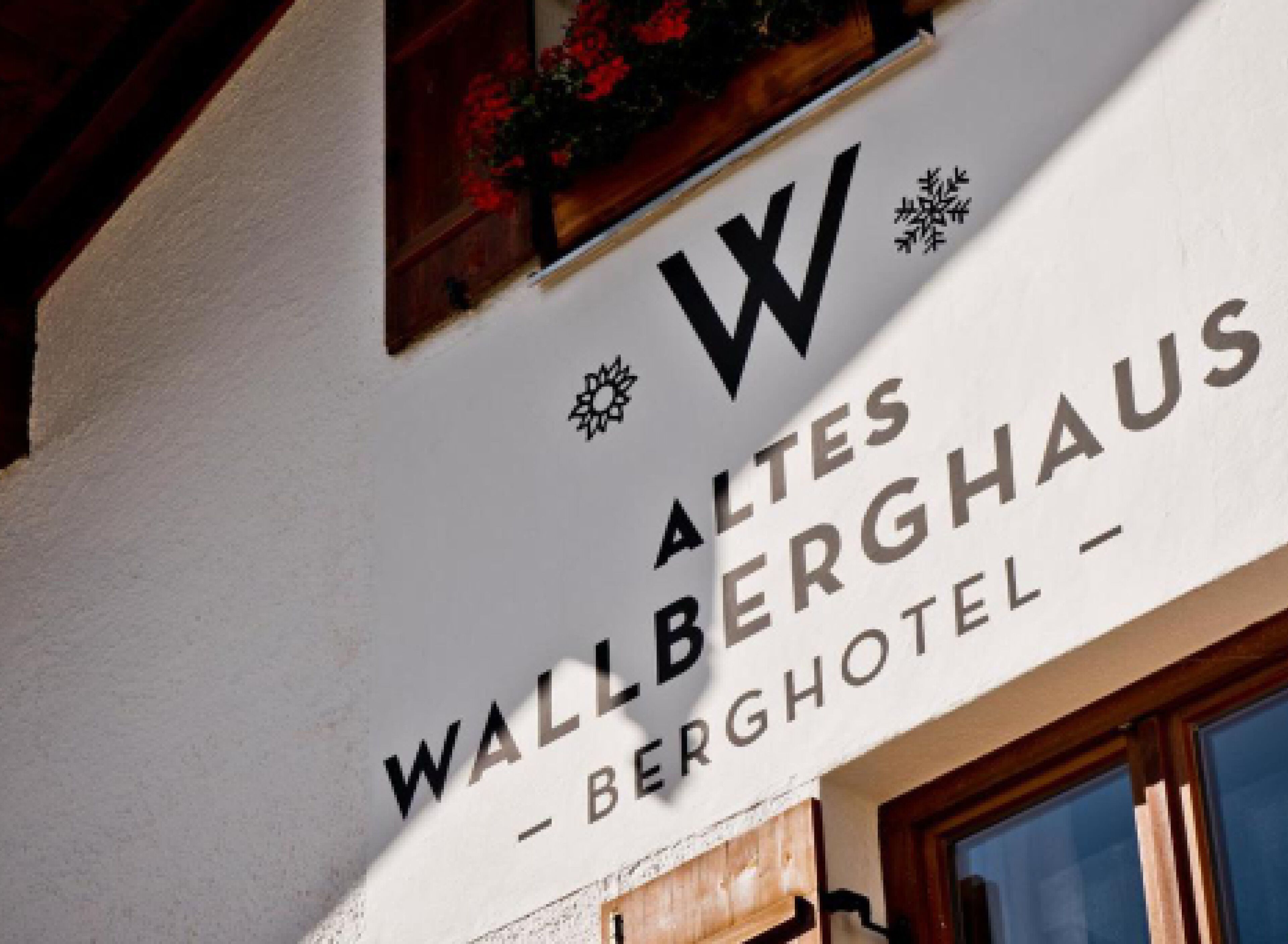 Ein Schild für 'Altes Wallberghaus - Berghotel' mit einem gut sichtbaren 'W'-Logo, Schneeflockenverzierung und einem hölzernen Fensterrahmen.