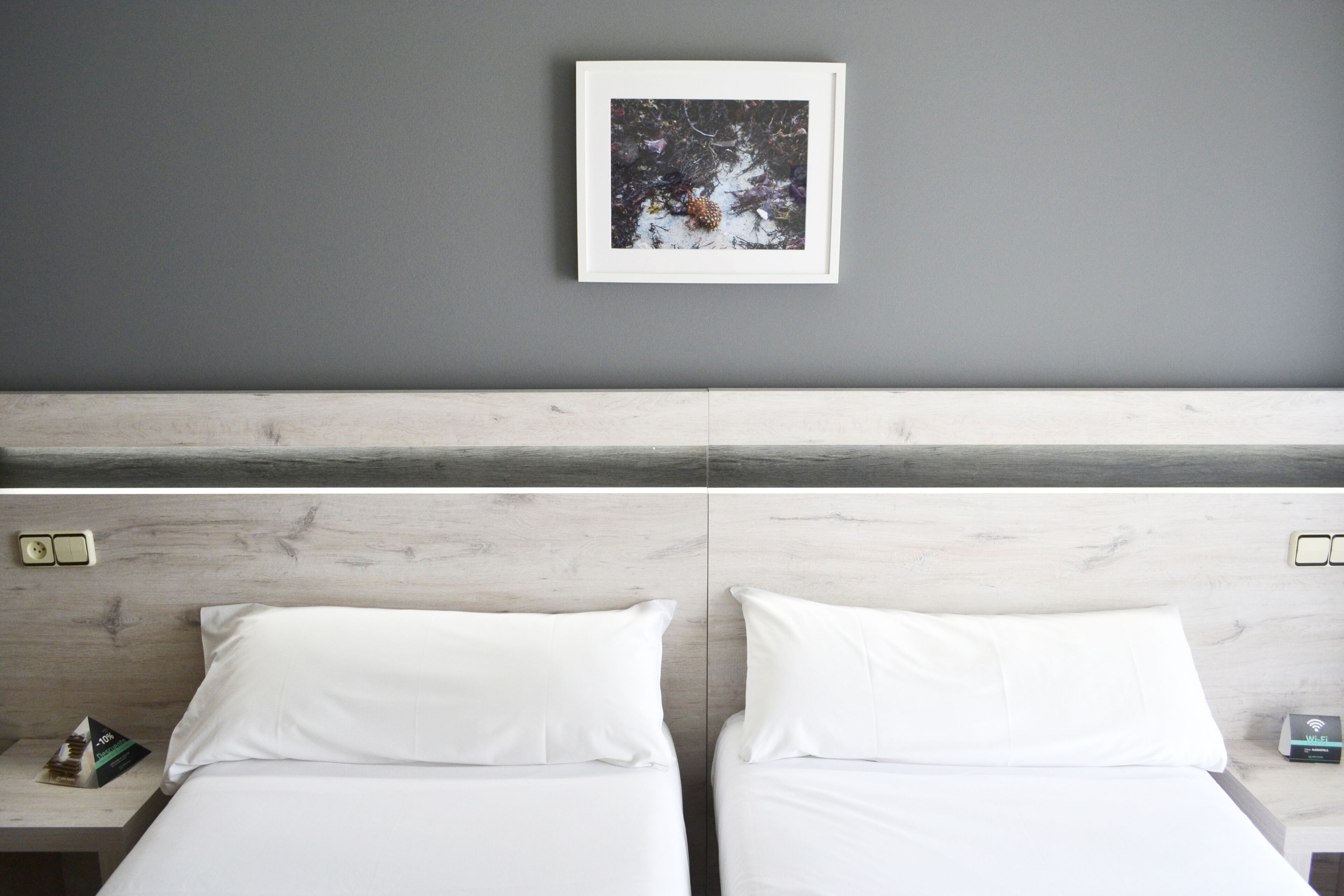 Una habitación de hotel con una pared gris, un cuadro enmarcado sobre la cama y un cabecero de madera.