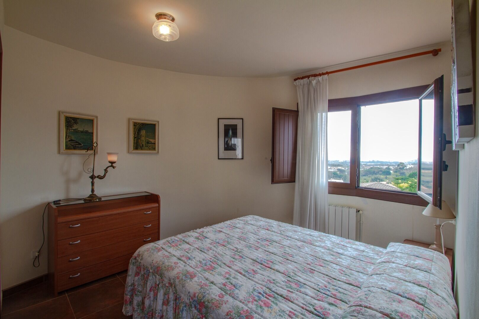 A bedroom with a bed, dresser, two framed pictures, a window with curtains, and a ceiling light.