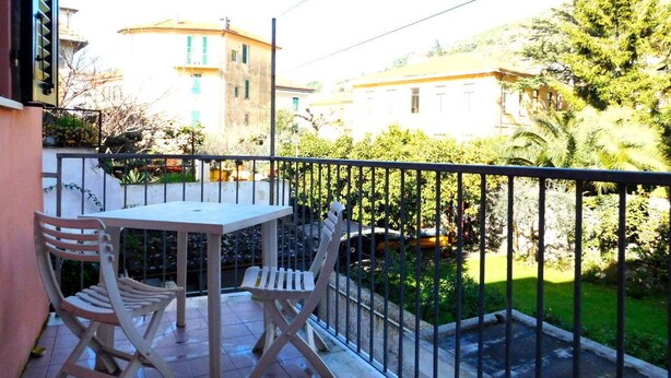 En balkon med et bord og to stole, der giver udsigt over en have og omkringliggende bygninger.