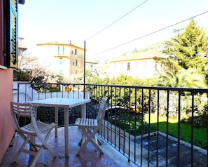 A balcony with a table and two chairs, overlooking a garden and neighboring buildings.