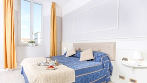 Une chambre à coucher bien rangée, avec un lit, une table de chevet, une fenêtre avec des rideaux et une vue sur les bâtiments à l’extérieur.