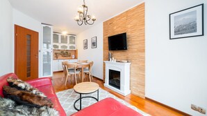 A modern living room with a fireplace, wooden wall paneling, a TV mounted above it, and a dining area with a kitchen in the background. - Apartamenty Sun & Snow Osiedle Podgórze (Szklarska Poreba)