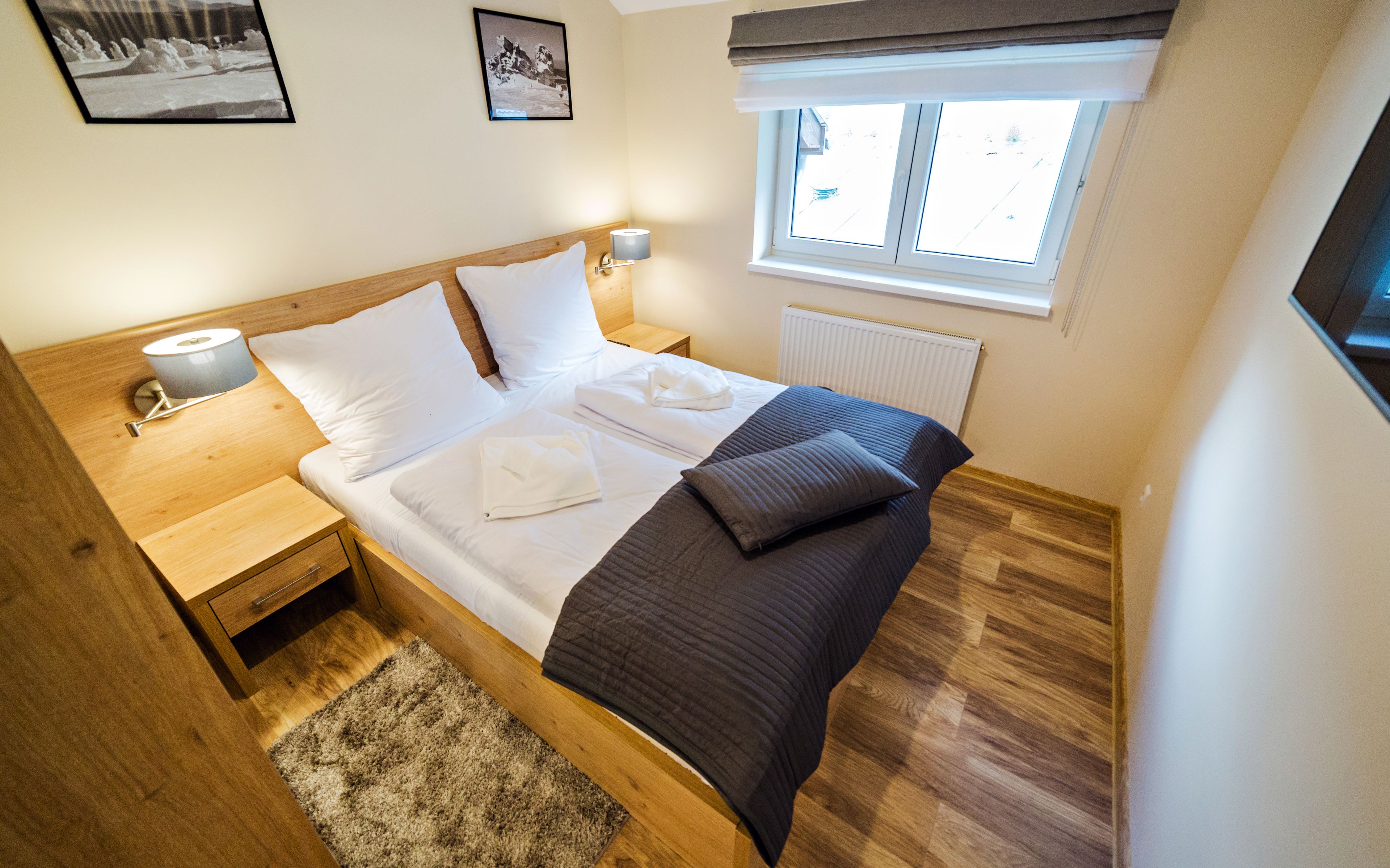 Ein ordentlich bezogenes Bett mit weißer und dunkler Bettwäsche, ein Nachttisch aus Holz mit einer Lampe, ein Heizkörper und ein Fenster mit Jalousien.