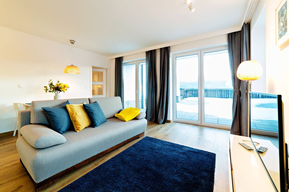 Ein modernes Wohnzimmer mit einem grauen Sofa, blauen und gelben Kissen, einem dunkelblauen Teppich und einem großen Fenster mit Vorhängen.