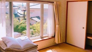 A room with tatami flooring, a window with curtains, a stack of pillows, and a wardrobe.