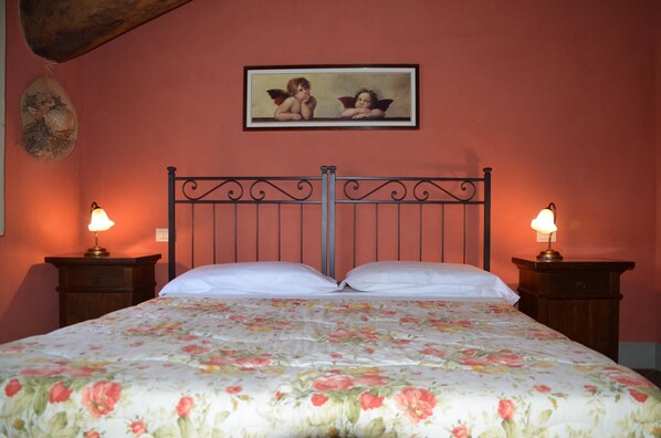 A bed with a floral bedspread, two bedside tables with lamps, a wall-mounted picture of cherubs, and a wooden ceiling beam.
