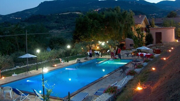 Una piscina con sillas de descanso y mesas, rodeada por un jardín ajardinado y vistas a una colina.