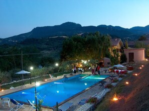 Una piscina con sillas de descanso y mesas, rodeada por un jardín ajardinado y vistas a una colina.