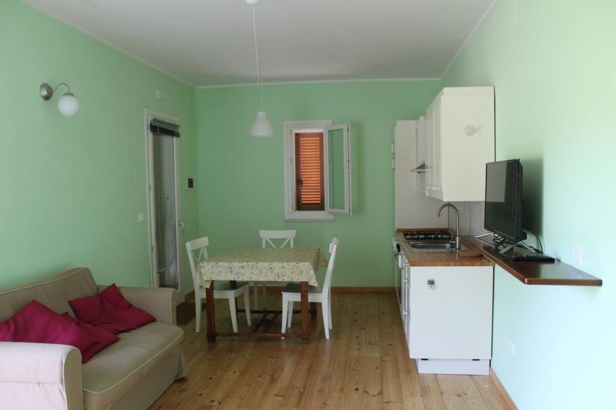 A small, well-lit room with a kitchenette, a dining area with a table and chairs, and a sofa with cushions.