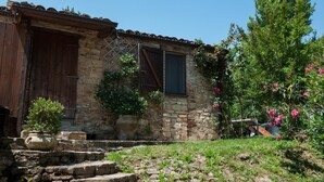 Edificio de piedra rústico con puerta y contraventanas de madera, rodeado por un jardín con plantas en flor.
