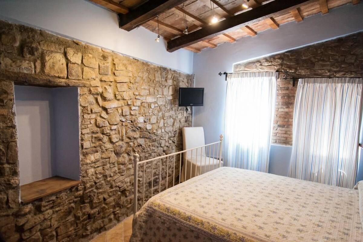 A bedroom with a stone wall, a bed, a TV mounted on the wall, and a window with curtains.