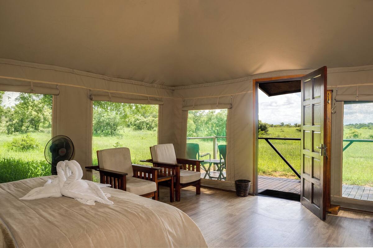 Ein Schlafzimmer mit einem Bett, zwei Stühlen, einem kleinen Tisch, einem Ventilator und einem Balkon mit Blick ins Grüne.
