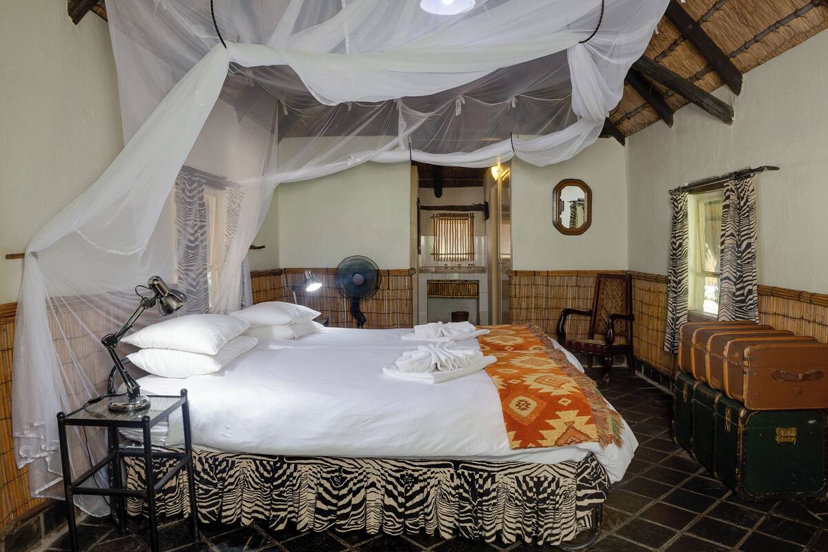 Ein Bett mit Baldachin, ein Nachttisch mit Lampe, ein Ventilator und ein Bettlaken mit Zeprazier. Koffer sind an der Wand gestapelt.