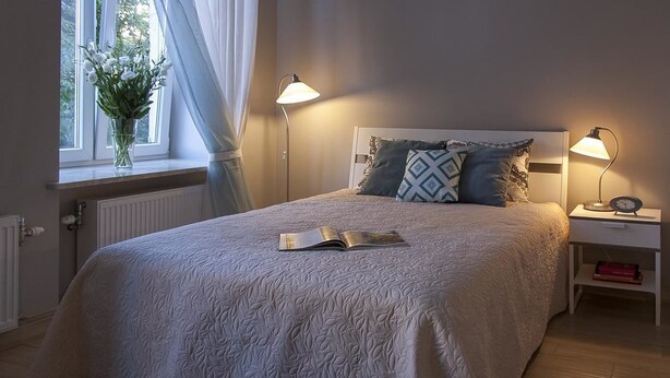 A bedroom with a large bed, two bedside lamps, a window with curtains, and a radiator. - Apartamenty 2M (Czestochowa)