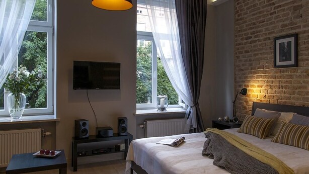 A bedroom with a large bed, a TV mounted on the wall, a brick wall, and a window with curtains. - Apartamenty 2M (Czestochowa)