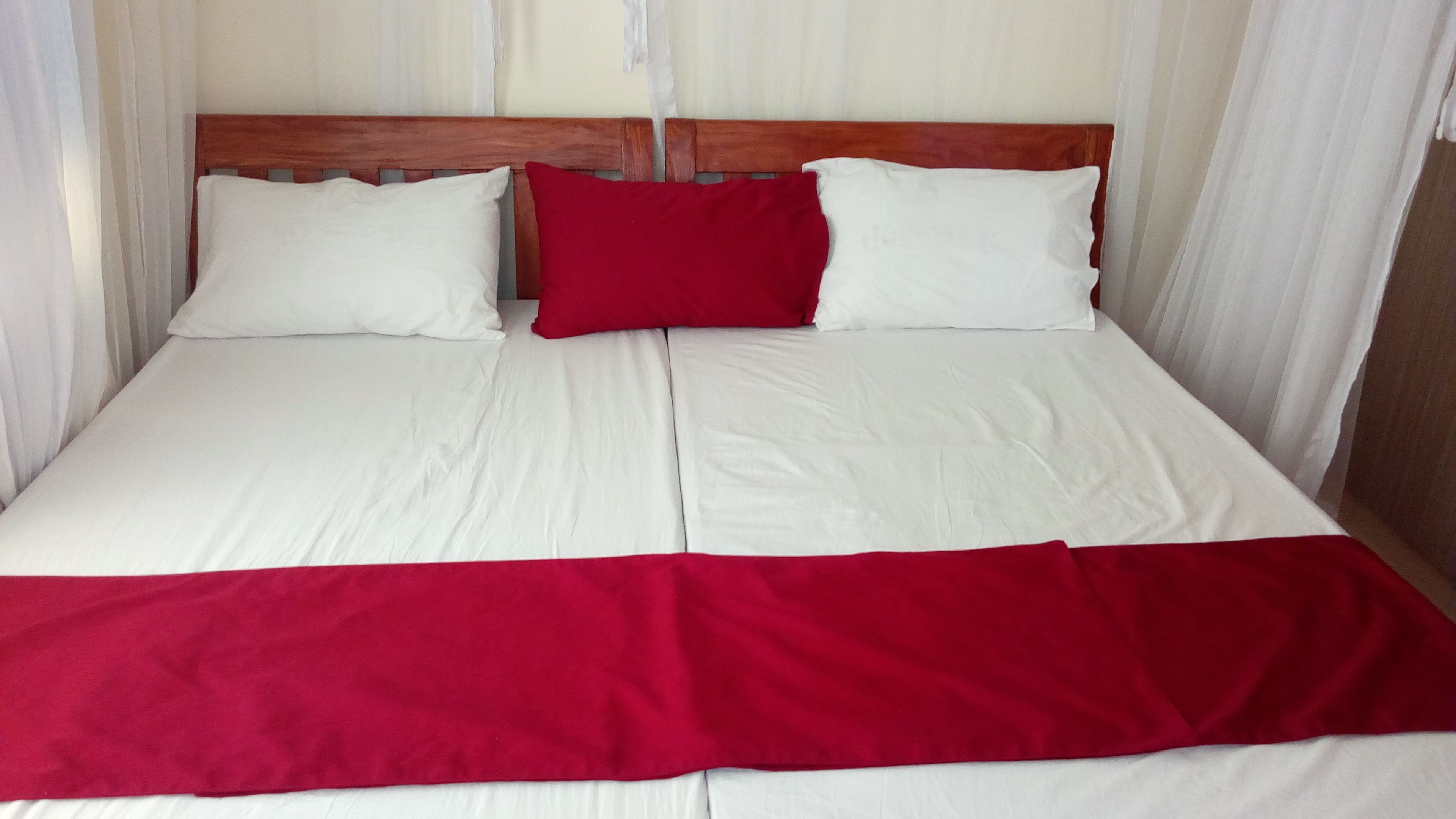 A bed with white bedding and a red accent pillow, a wooden headboard, and sheer curtains.