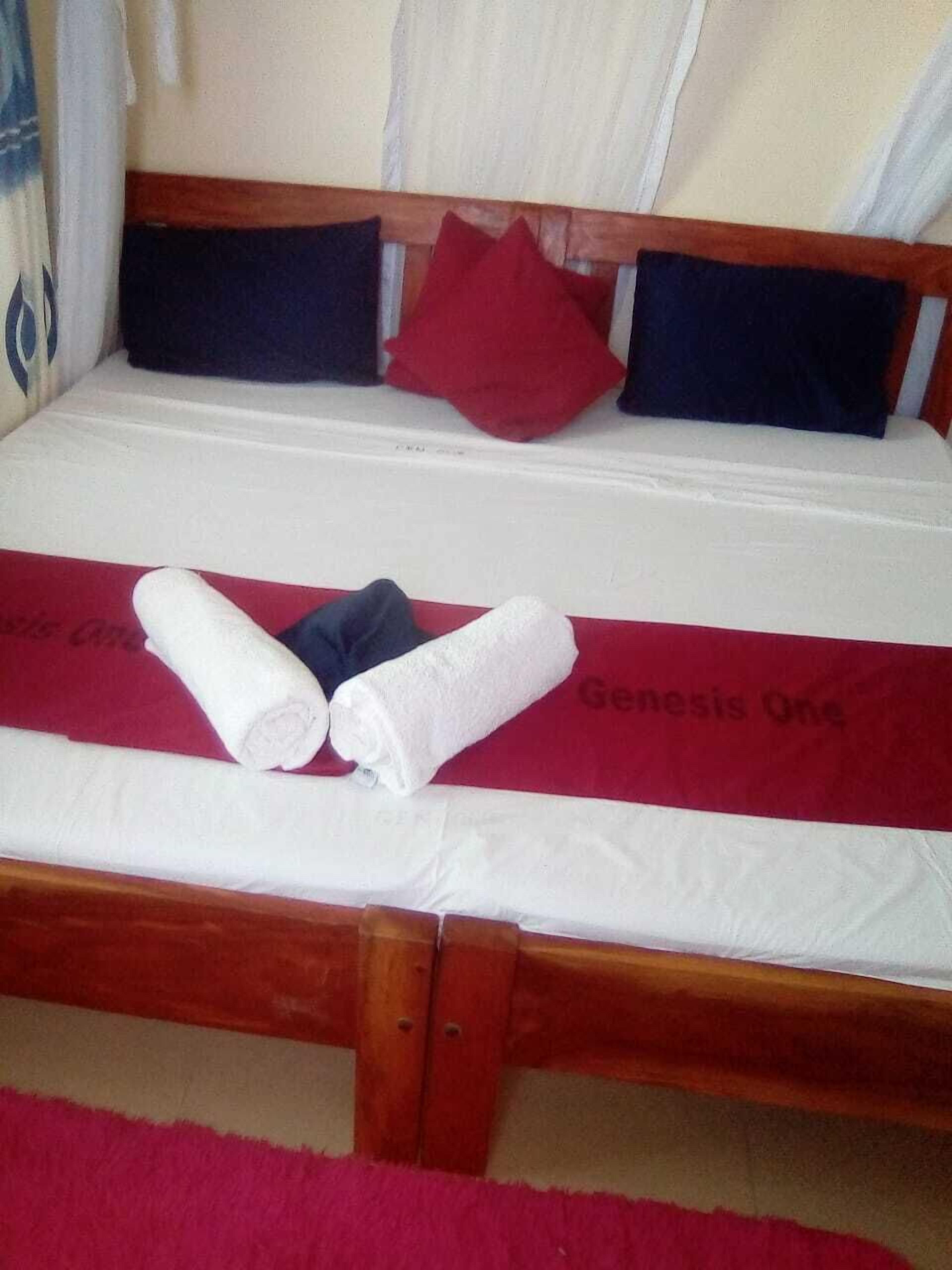 Un lit avec un matelas blanc et une couverture rouge sur laquelle est imprimé le texte « Genesis One ». Deux serviettes blanches enroulées sont posées sur le lit.