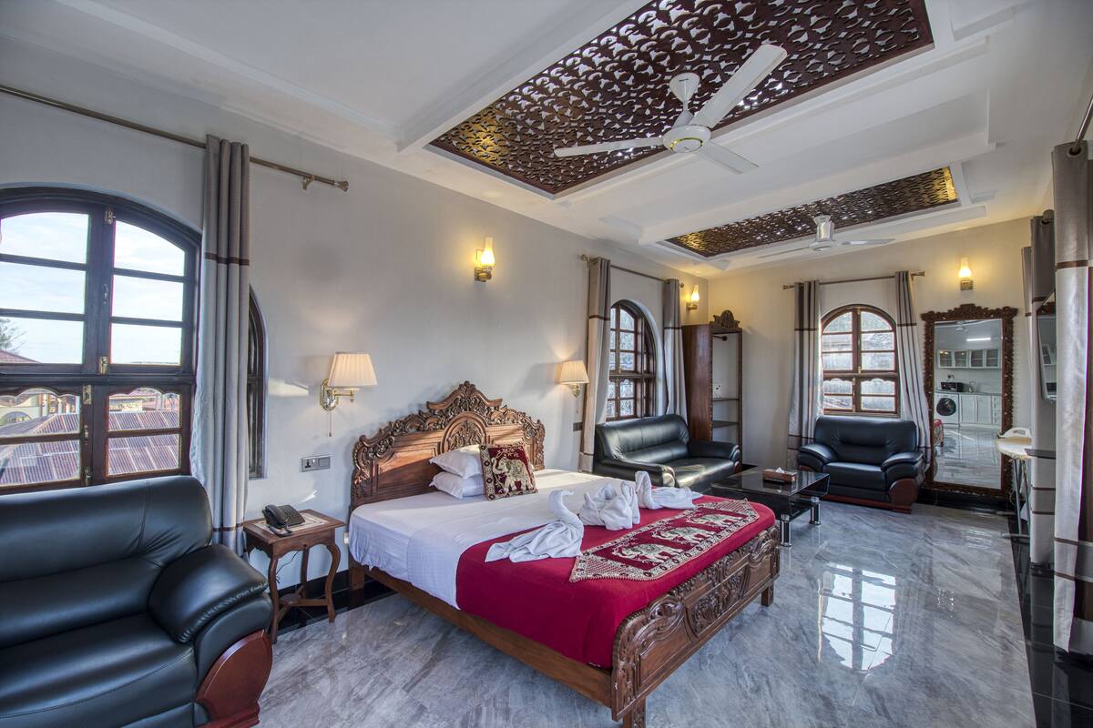 A hotel room with a large bed, two black leather chairs, a wooden headboard, a ceiling fan, and a view of the outside through a large window.