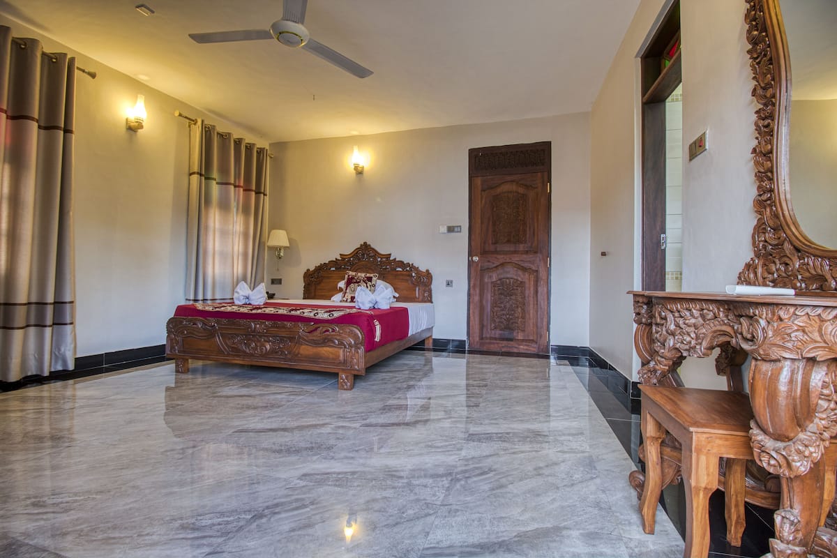 A spacious hotel room with a large bed, a wooden headboard, a ceiling fan, and a wooden side table.