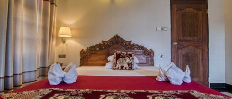 A hotel room with a large bed, a wooden headboard, a red and white patterned bedspread, a wall-mounted lamp, and a door with intricate carvings.