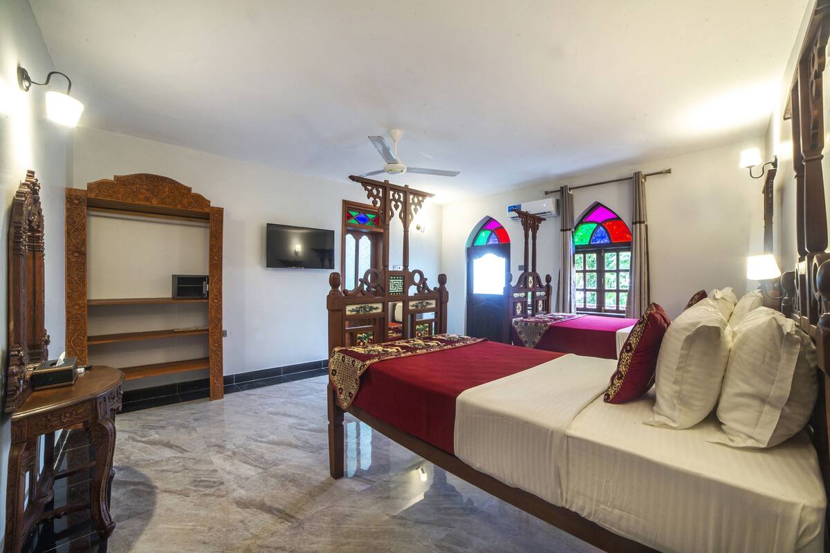 A bedroom with a bed, a ceiling fan, a wooden chair, a table, and a TV on the wall.