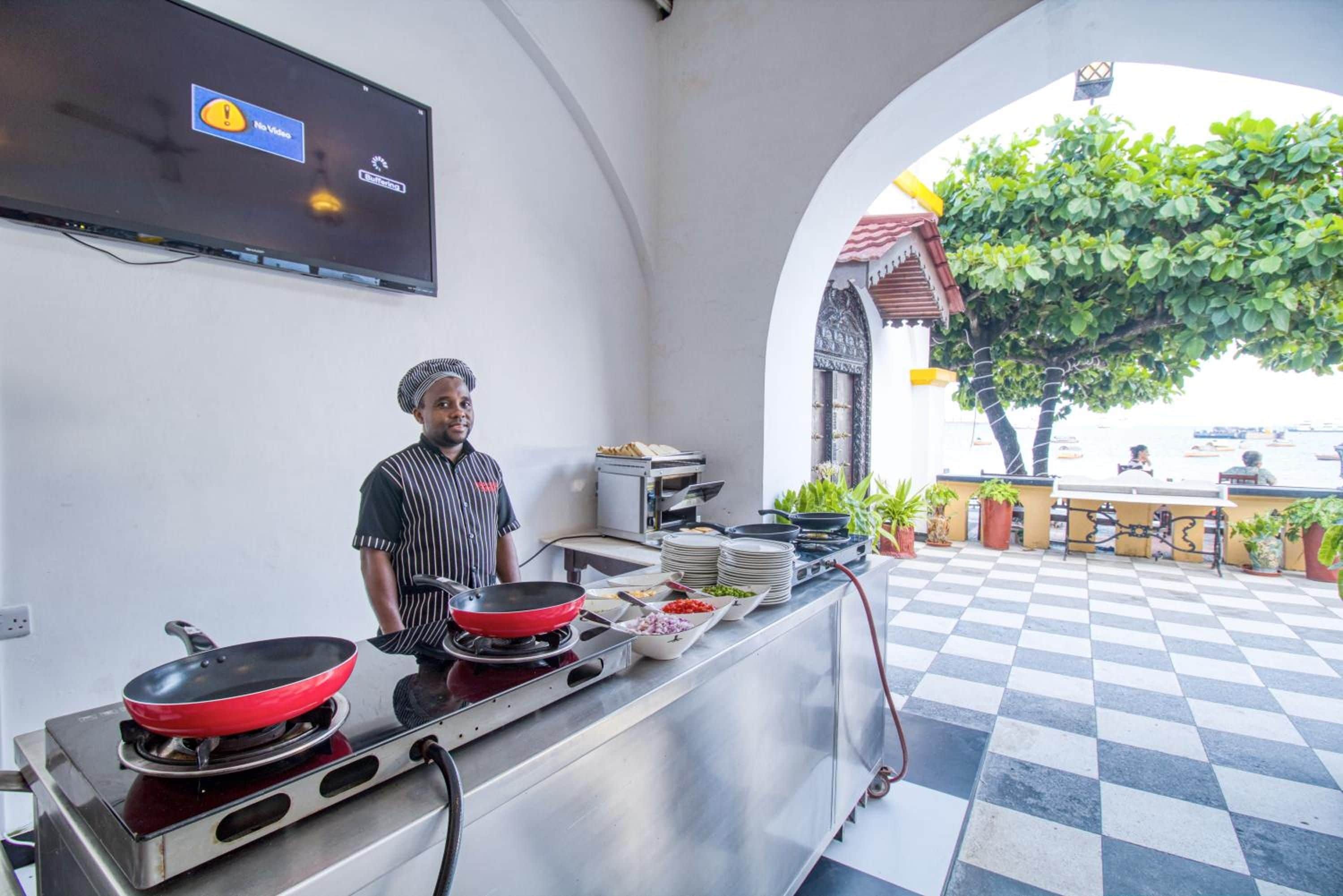 A chef in a kitchen with a TV displaying a buffering screen, a checkered floor, and a view of a tree and building outside.