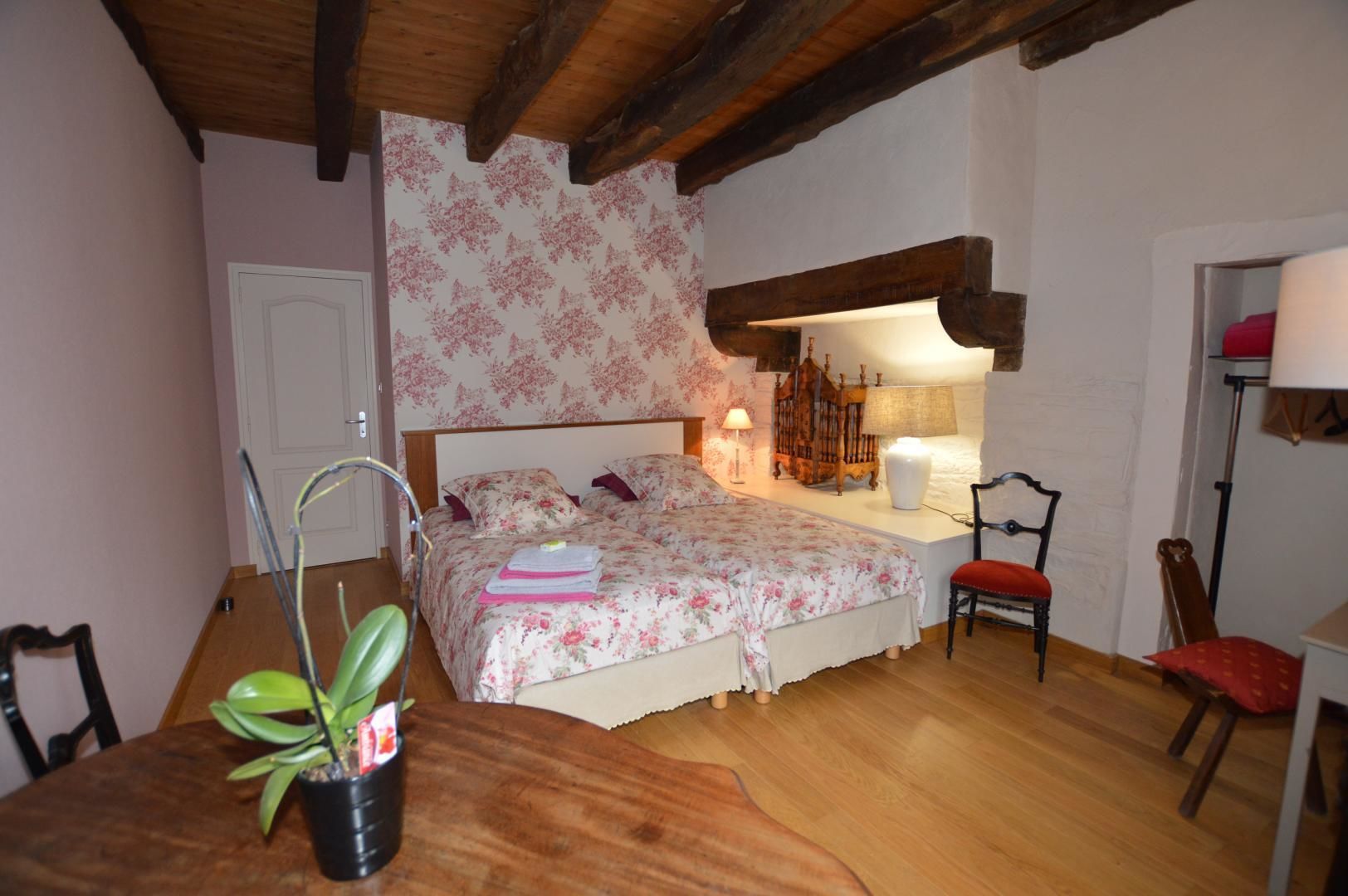 A bedroom with a wooden ceiling, floral wallpaper, a bed with floral bedding, a bedside table with a lamp, a chair, and a small table with a plant.