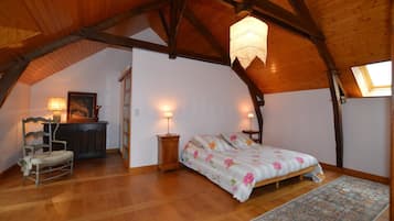 A cozy attic bedroom with a wooden ceiling, a bed with floral bedding, a chair, a desk with a lamp, and a painting on the wall.