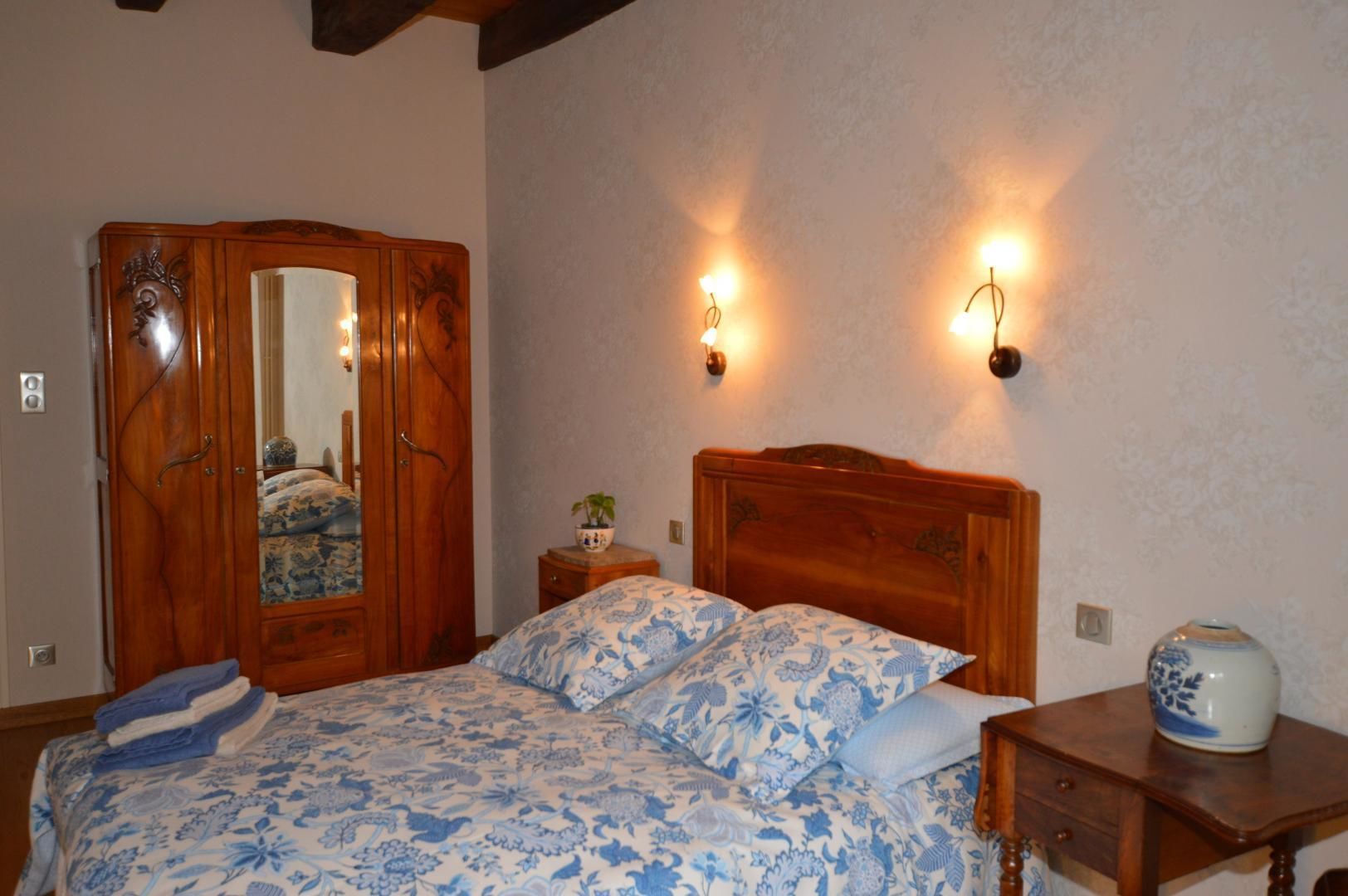 A bedroom with a wooden bed, a wardrobe, two wall-mounted lamps, and a small bedside table with a vase.