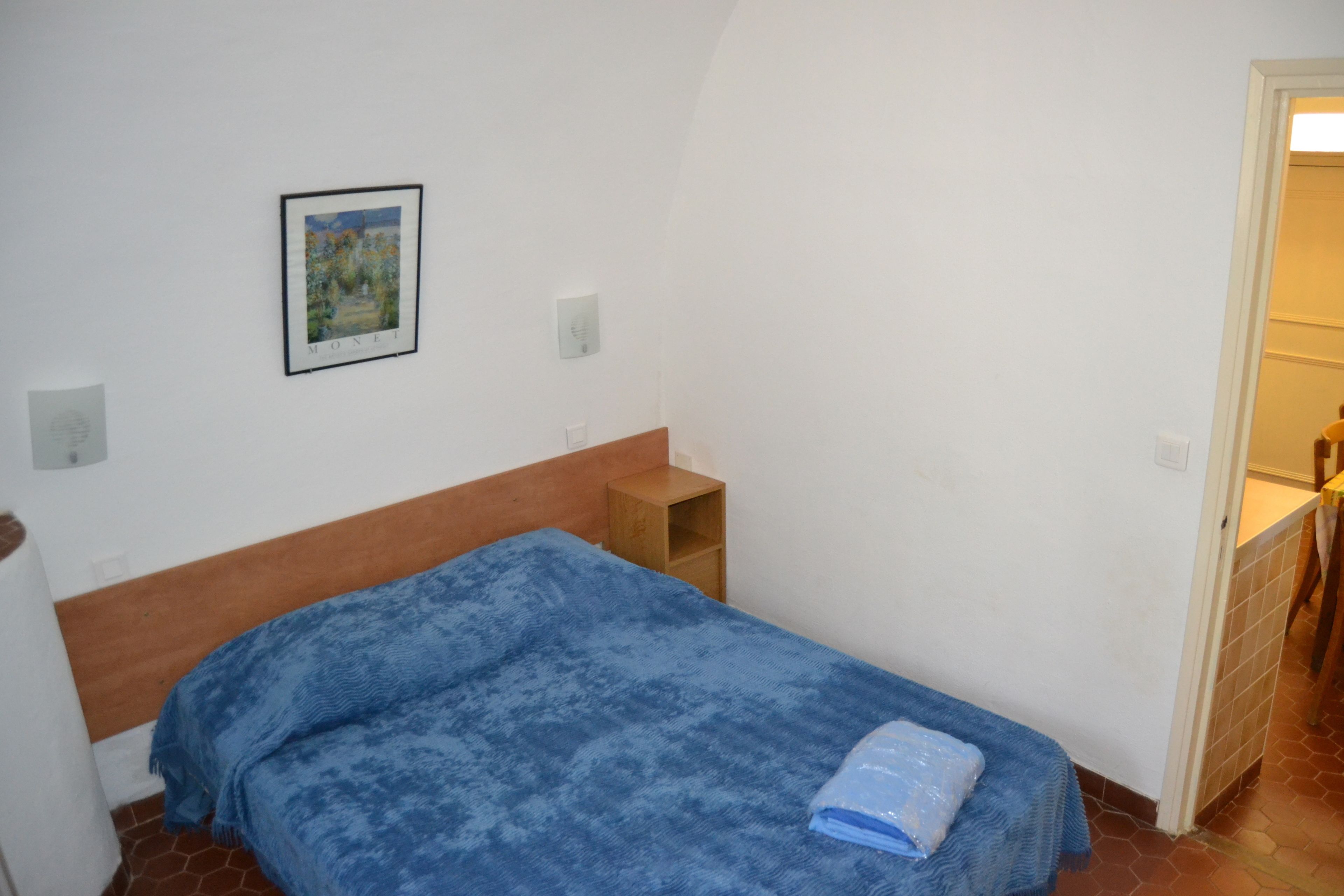 Una habitación pequeña y sencilla con una cama individual, una mesita de noche de madera y un cuadro en la pared.