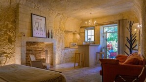 A rustic stone room with a bed, a red armchair, a fireplace, and a window with a view of greenery outside. - Le Gaimont (Vouvray)