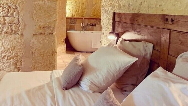 A stone-walled bedroom with a wooden headboard, a bed with white linens, and a bathtub in the background. - Le Gaimont (Vouvray)