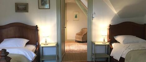 A bedroom with two beds, a wooden headboard, a nightstand with a lamp, a chair in the background, and a door leading to another room.