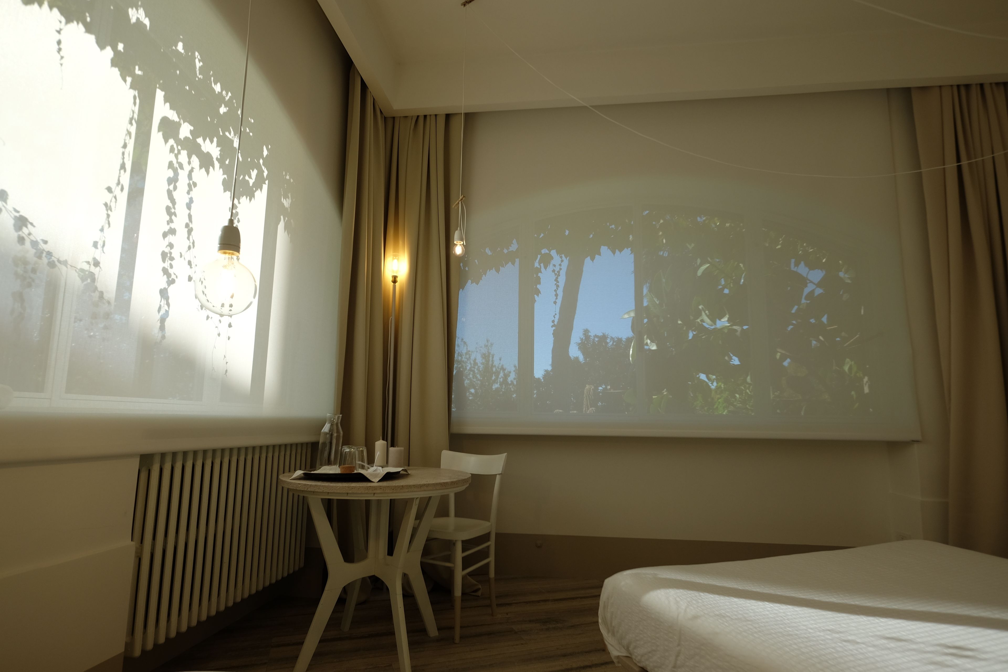 A hotel room with a bed, a small table with a chair, a projector screen, and a window with curtains.