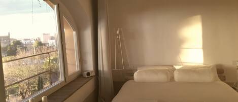 Un dormitorio con un ventanal grande, una cama con ropa de cama blanca, una mesita de noche y una lámpara.