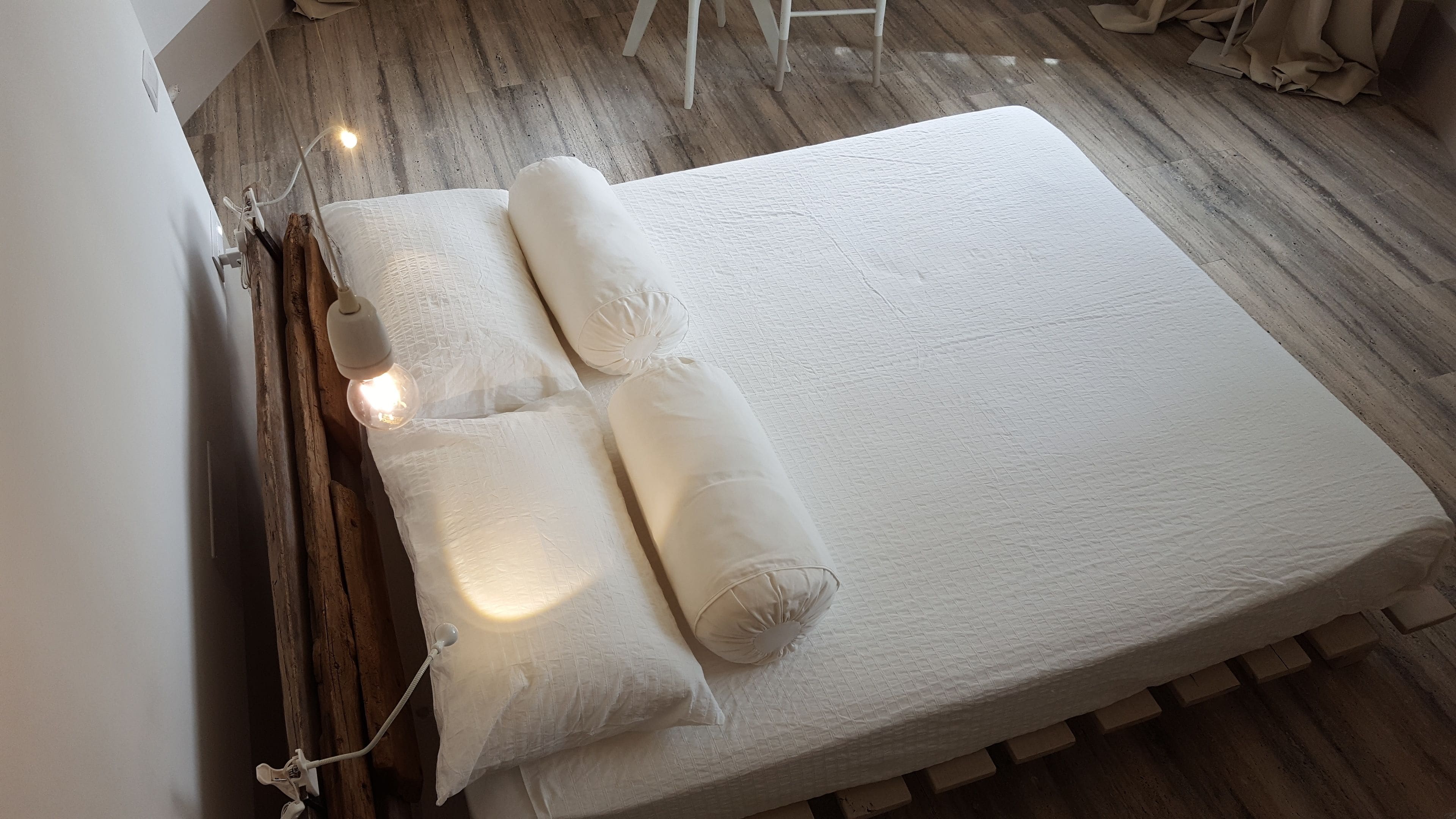 A neatly made bed with white linens and two rolled-up pillows, a bedside lamp, and a wooden floor.