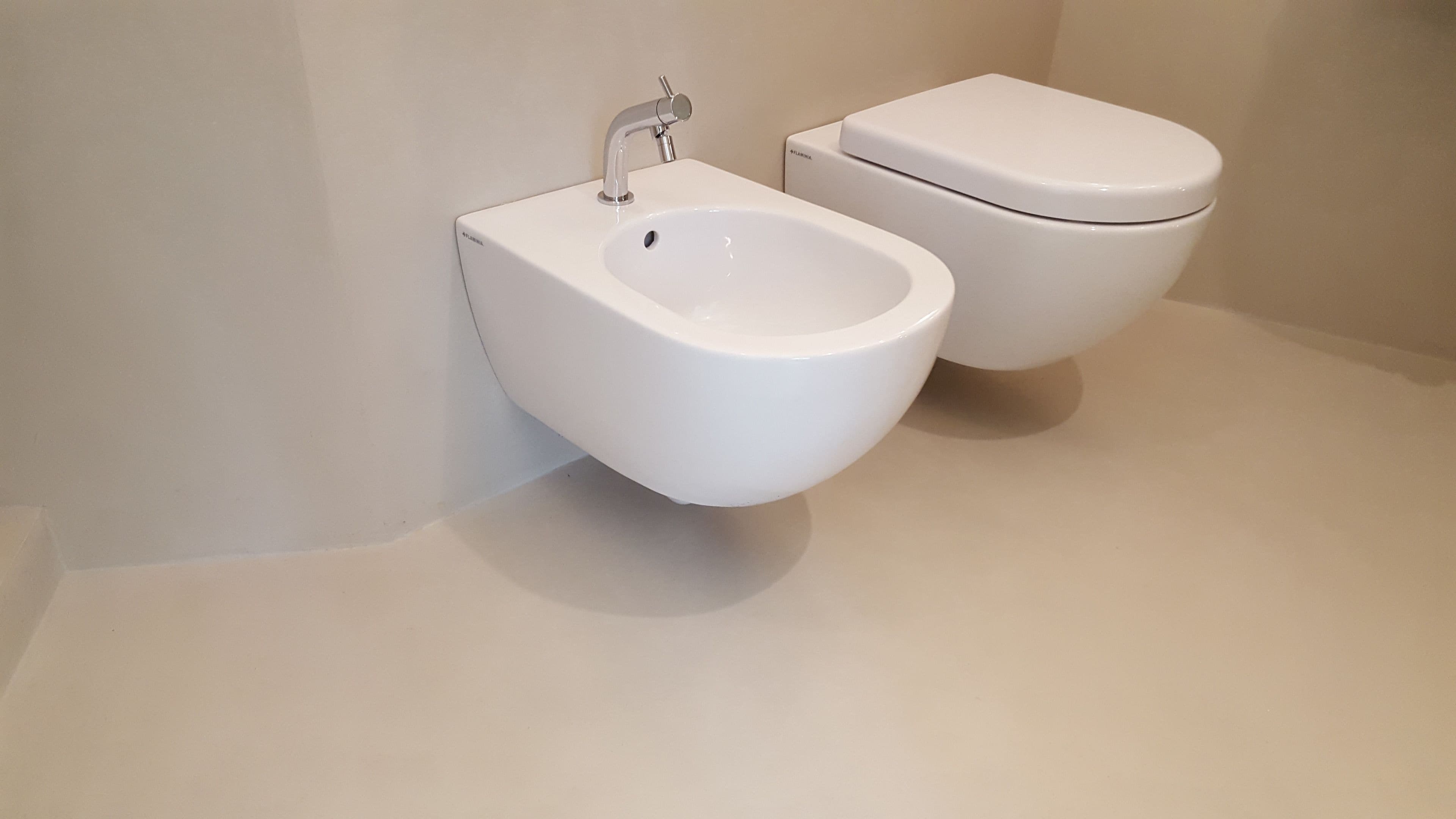 Ein weißes Bidet und eine Toilette in einem Badezimmer mit schlichter Wand und glattem Boden.