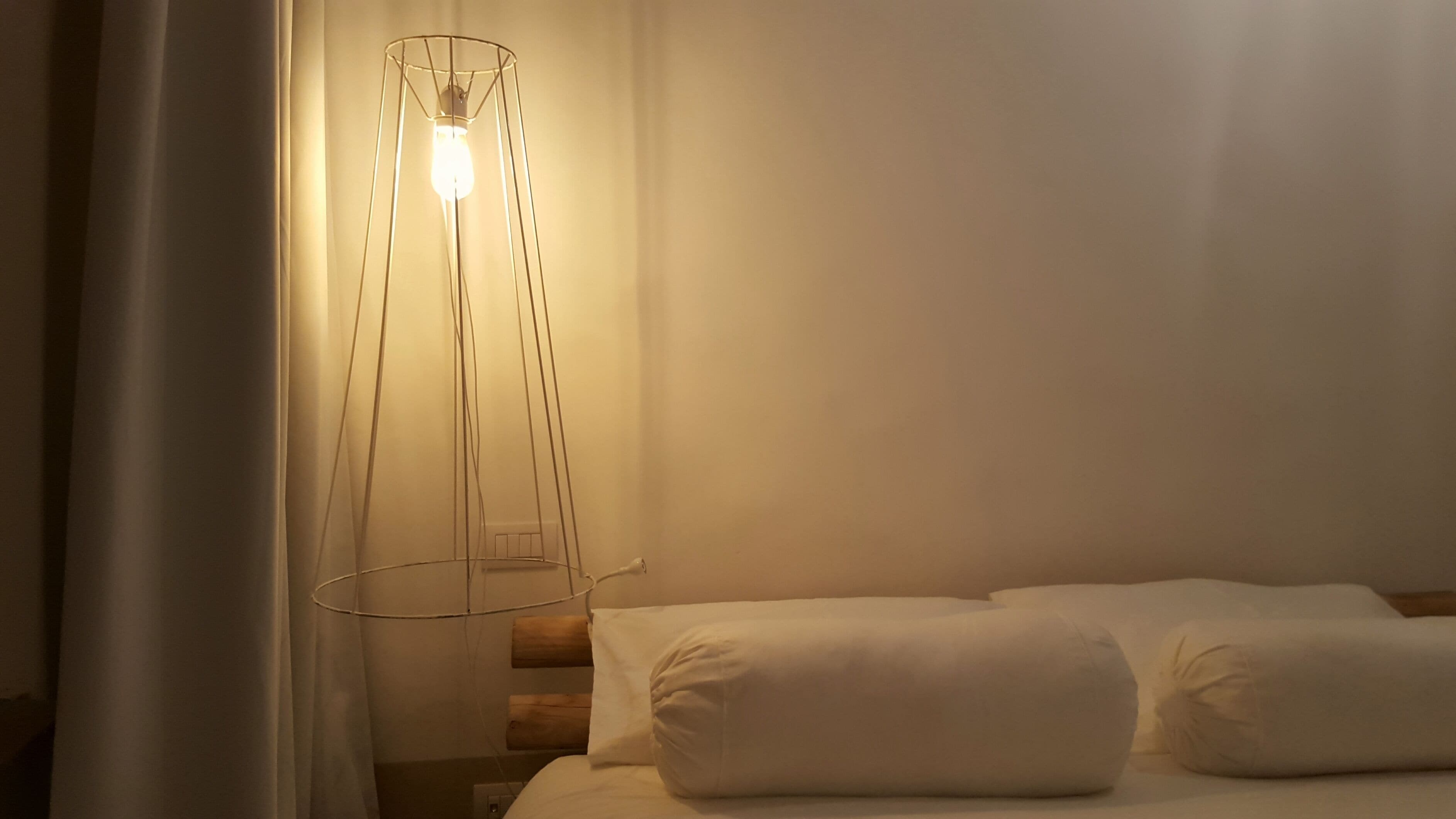 Una cama con sábanas blancas y una lámpara de pie moderna con una bombilla.