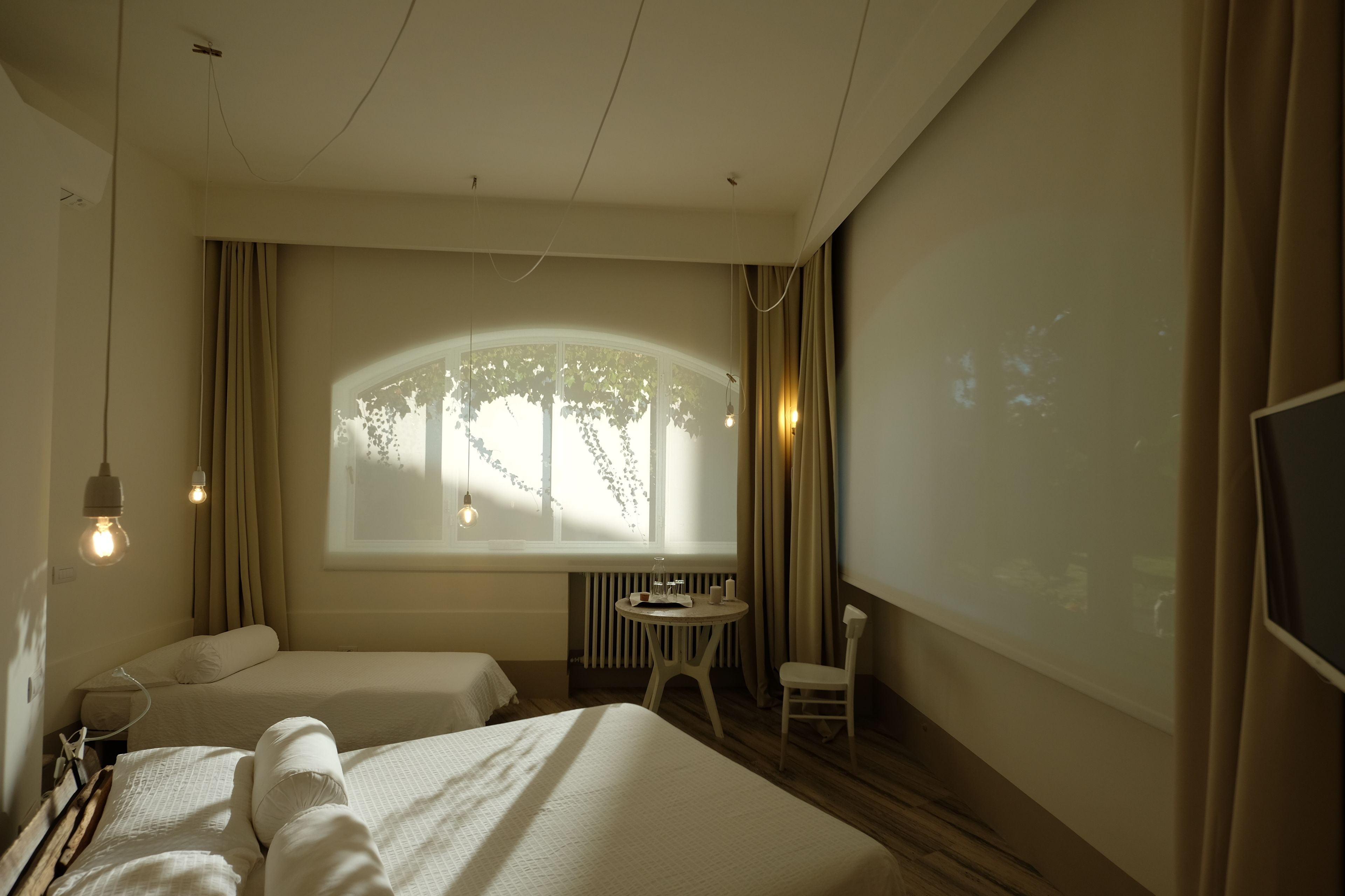 A hotel room with two beds, a small table, a chair, a window with curtains, and a projector screen.