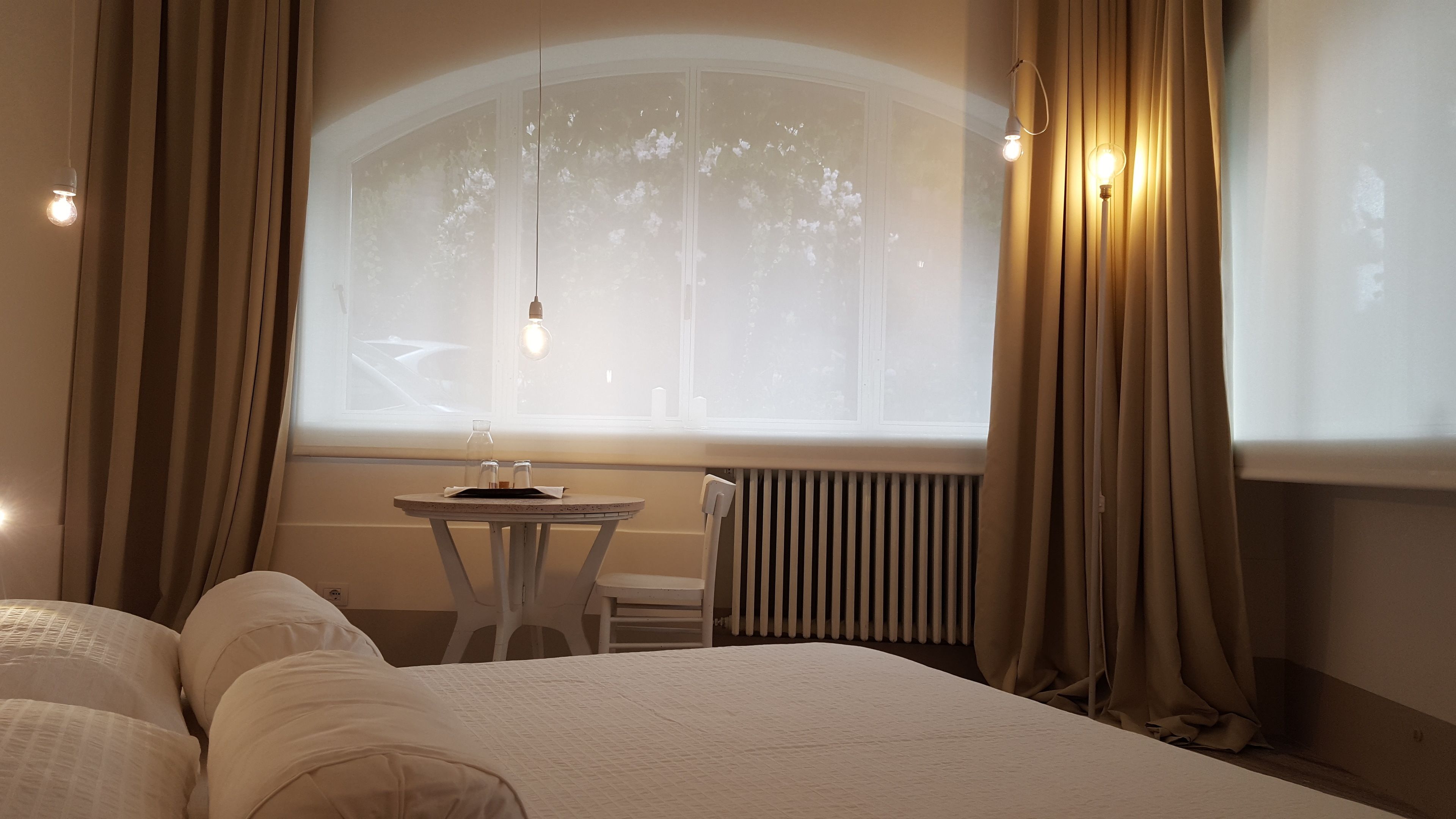 A hotel room with a large bed, a small table with a chair, a window with curtains, and a radiator.