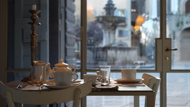 A table set with white cups, a glass, and a teapot, with a view of a fountain outside. - B&B A Piazza del Gesù (Viterbo)