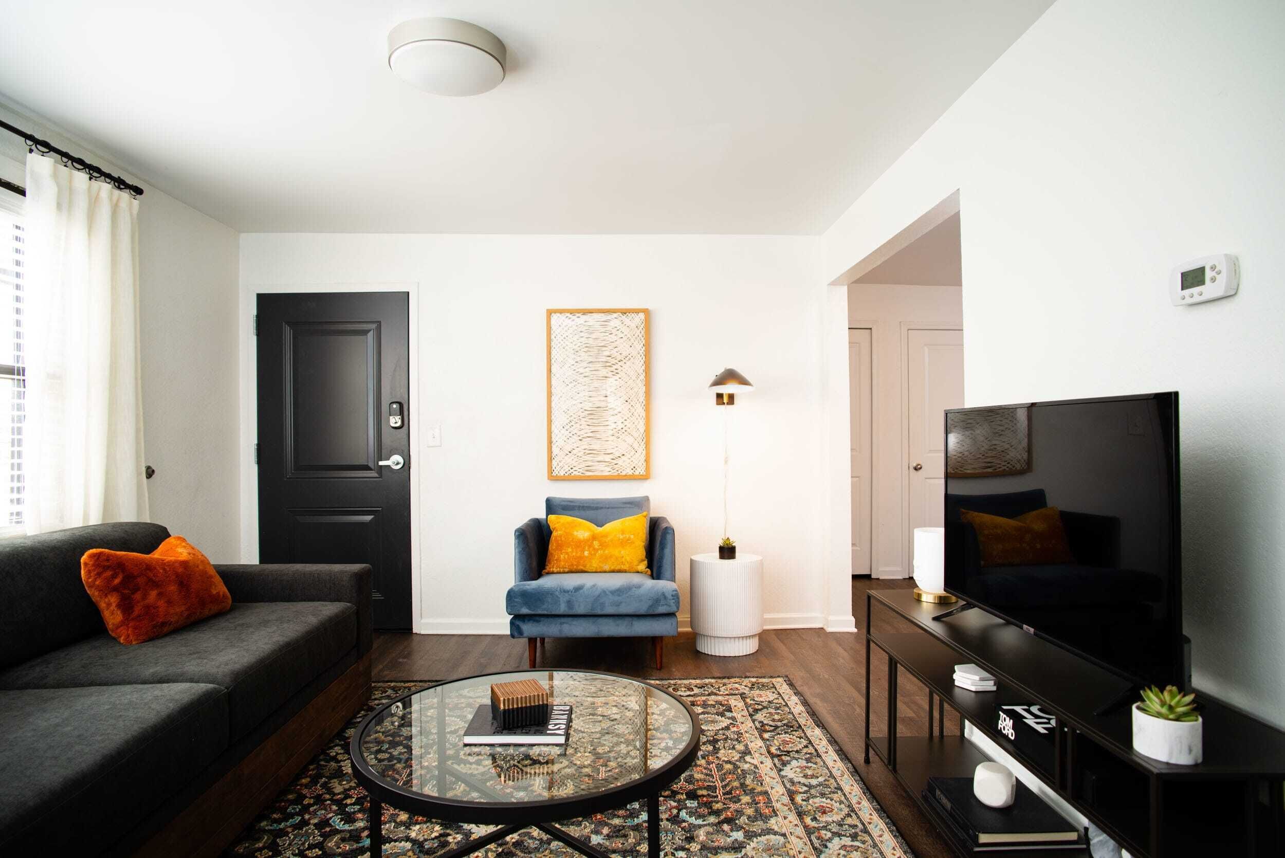 A modern living room with a dark sofa, a blue armchair, a round coffee table, a flat-screen TV, and a patterned rug.