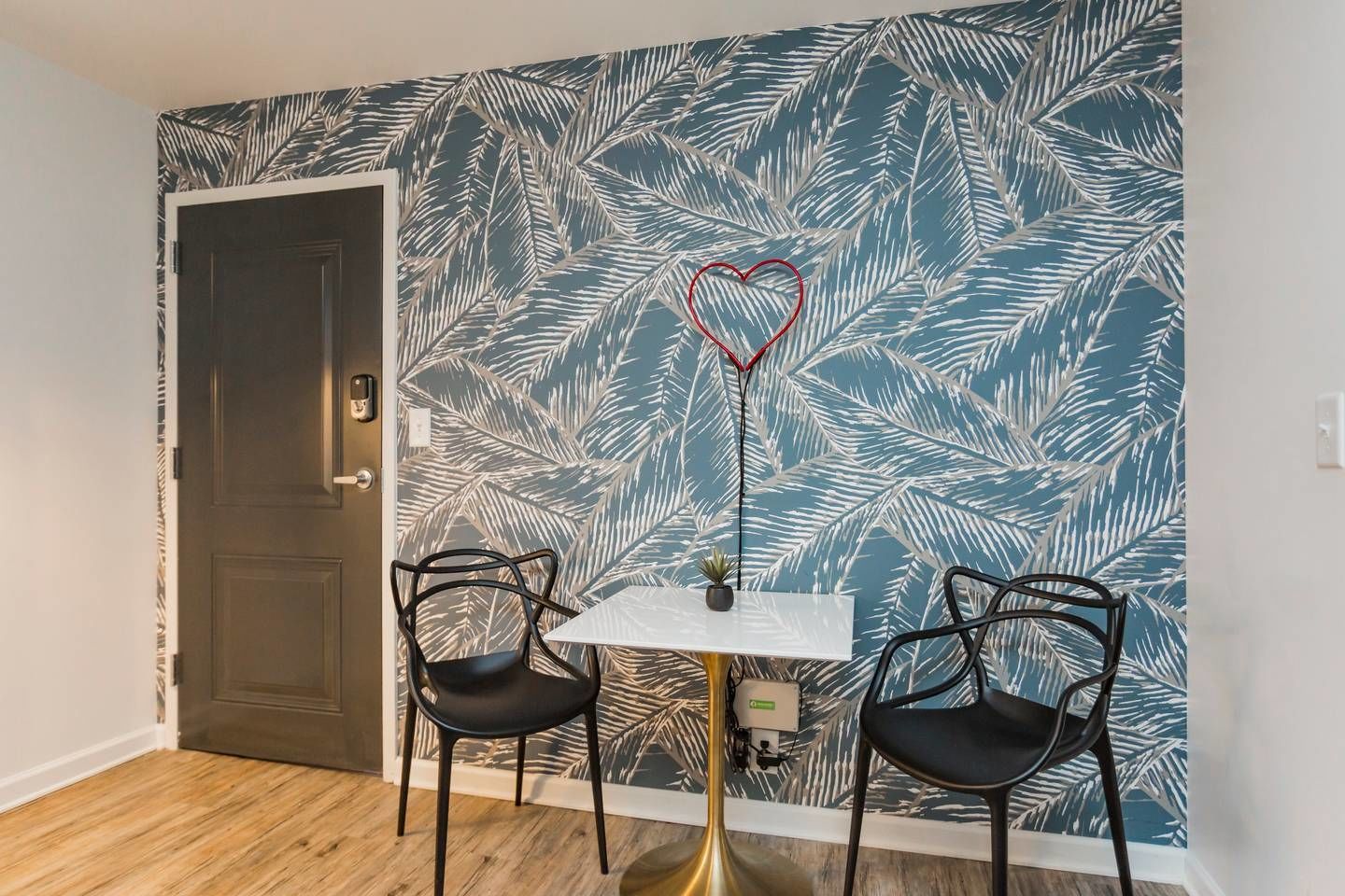 Una habitación con una puerta de madera oscura, una mesa blanca y dos sillas negras, y una pared con un diseño de hojas tropicales.