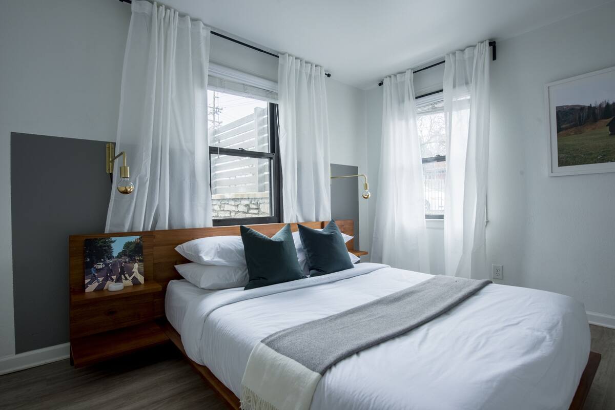 Een moderne slaapkamer met een groot bed, witte gordijnen en een schilderij op het nachtkastje.