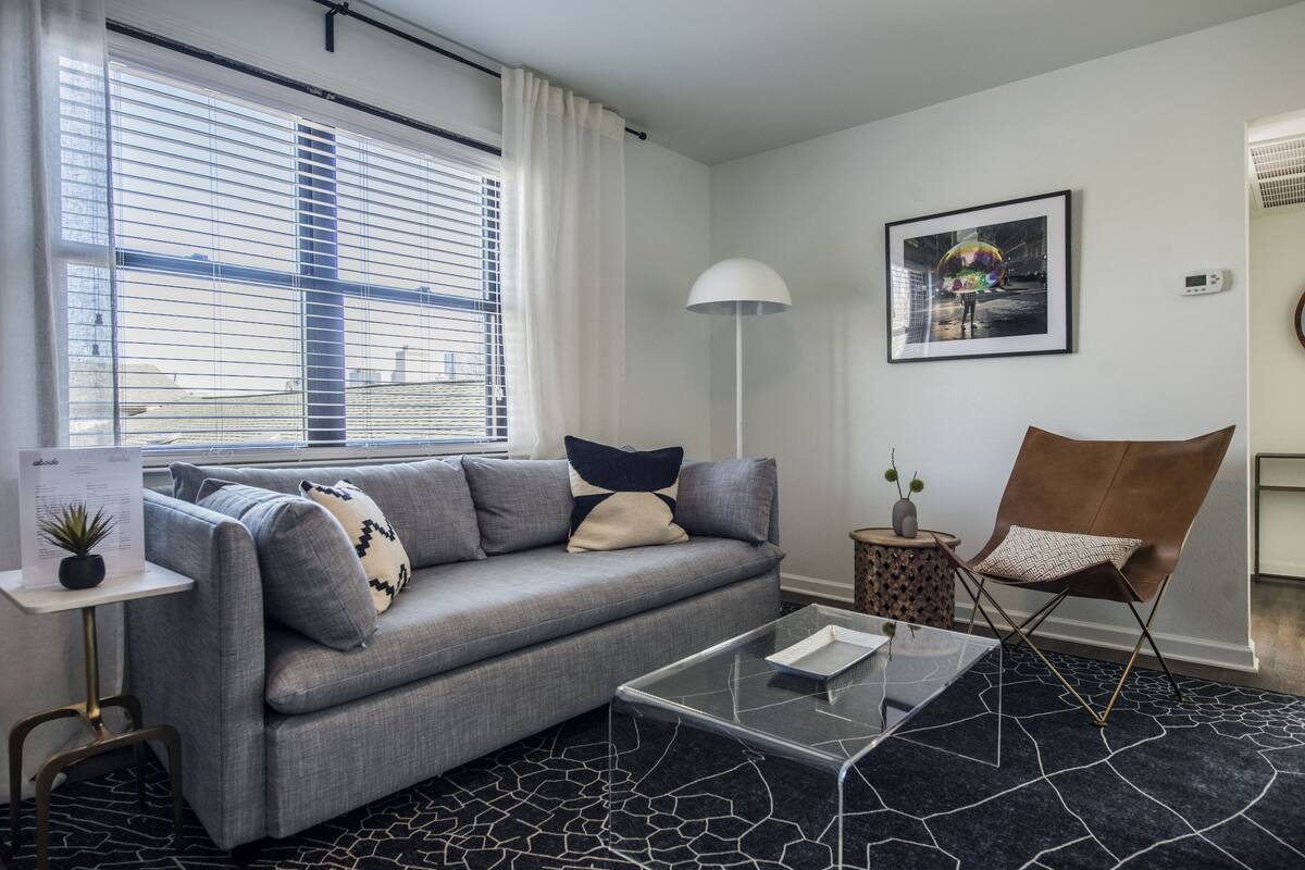 Een moderne woonkamer met een grijze bank, een glazen salontafel, een bruine fauteuil, een vloerlamp en een lijst met een foto aan de muur.
