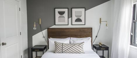 Un dormitorio ordenado con una cama grande, dos mesitas de noche, un cabecero y dos lámparas fijadas a la pared.
