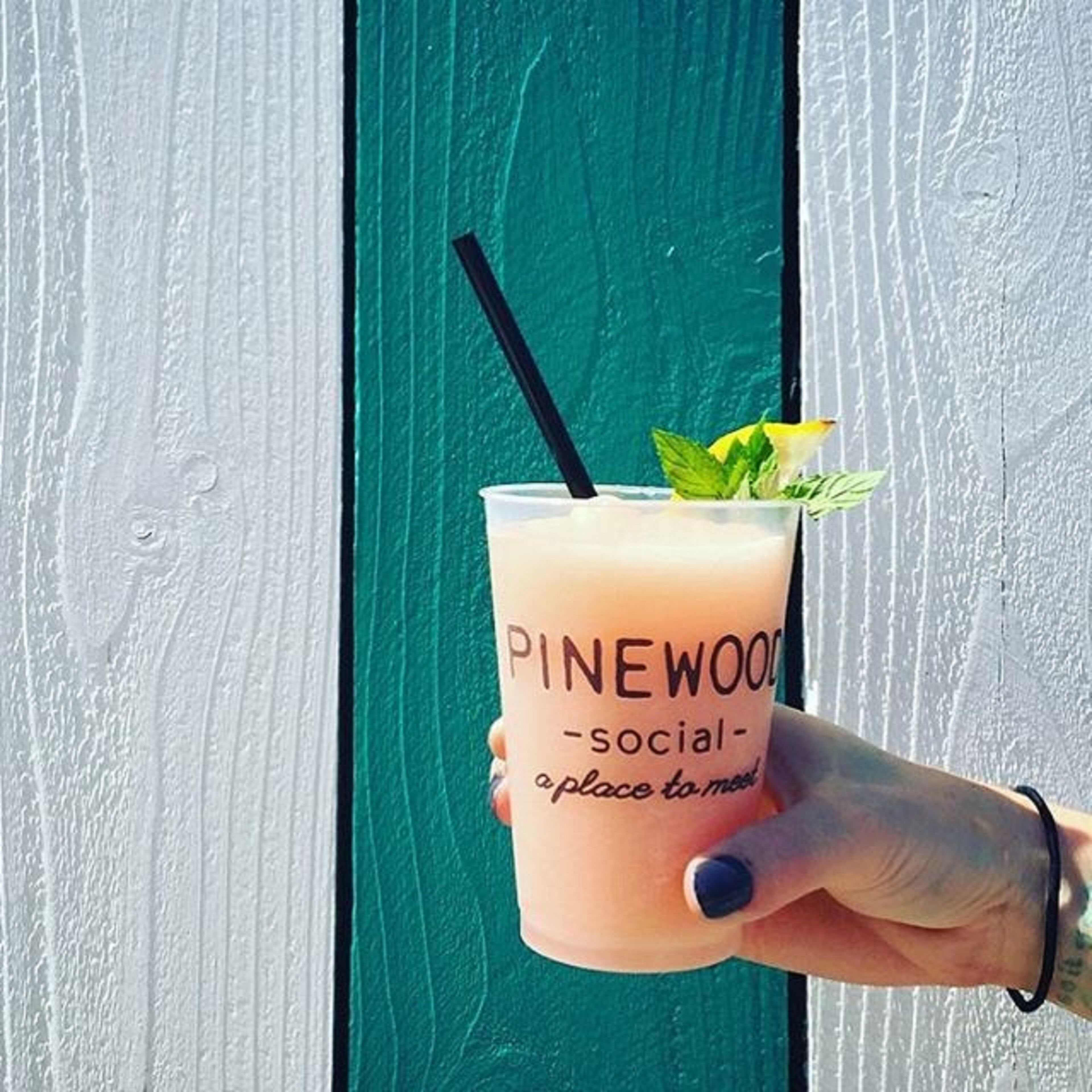 A hand holding a Pinewood Social - a place to meet drink with a black straw and mint garnish.